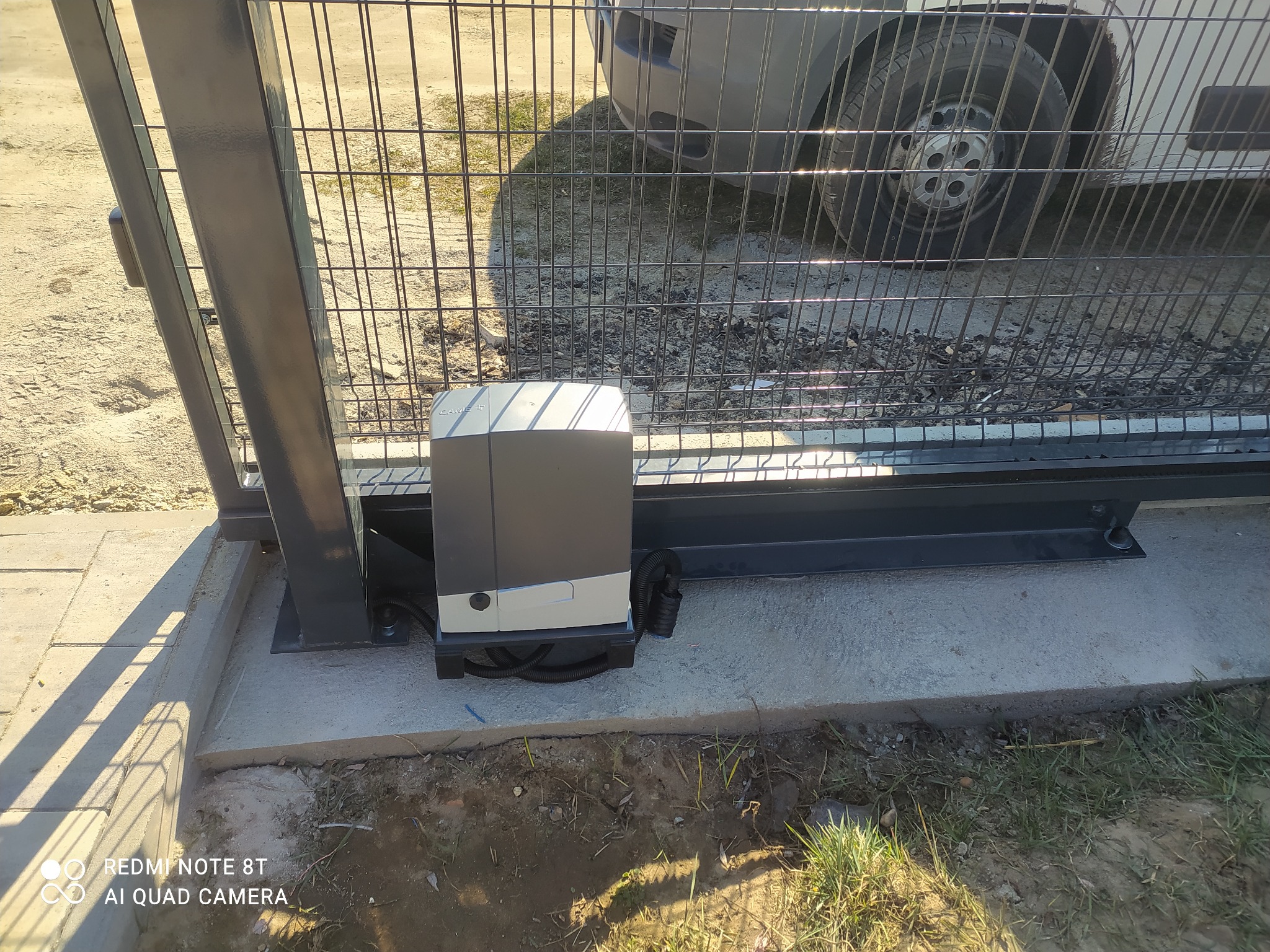 Szara brama przesuwna z automatyką, widoczny mechanizm napędowy zamontowany na betonowej podstawie, w tle fragment ogrodzenia z siatki i koło samochodu.