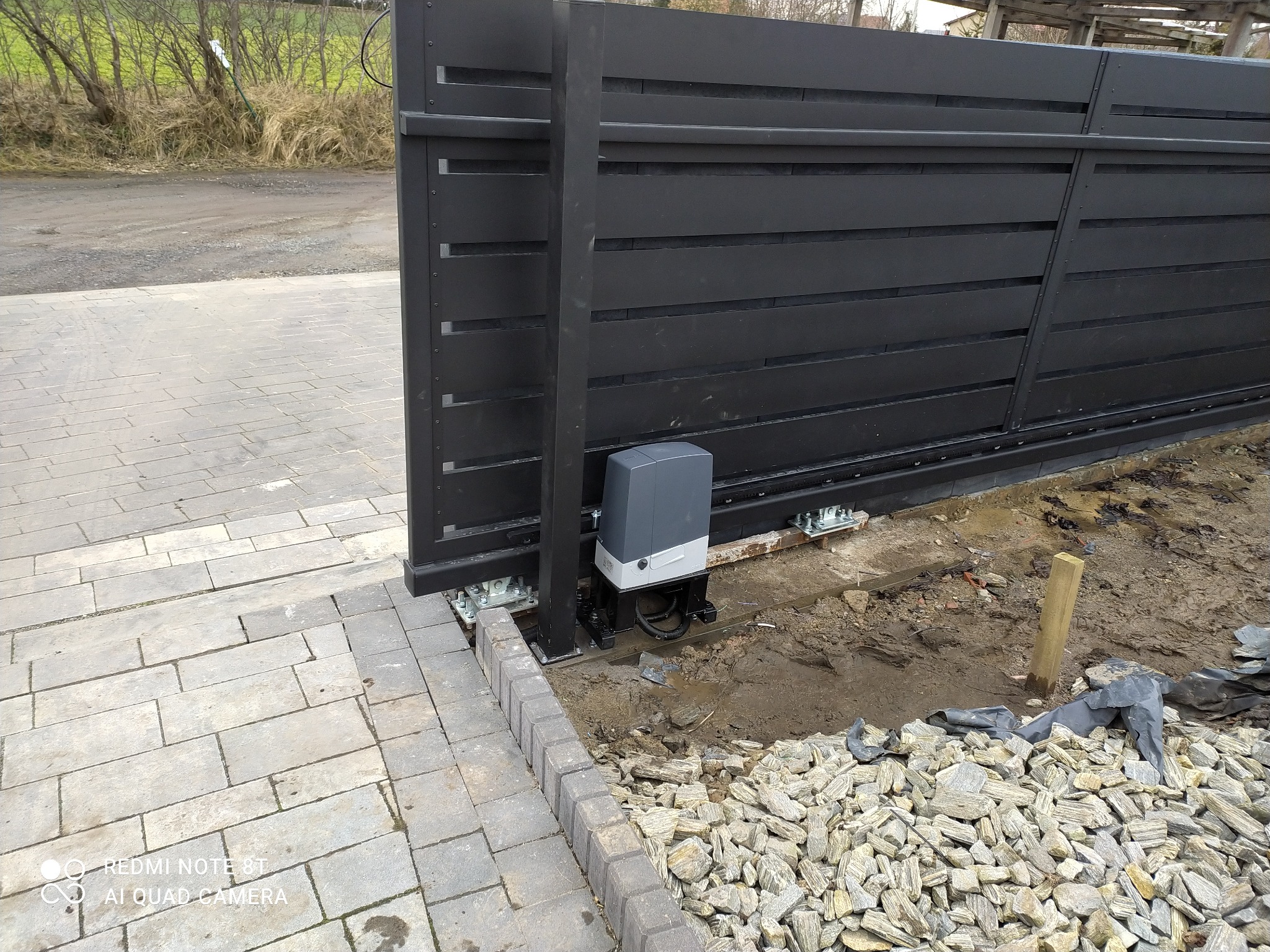 Czarna, przesuwna brama wjazdowa z automatycznym napędem, widoczny mechanizm i fundament w trakcie prac wykończeniowych z kamieniami.