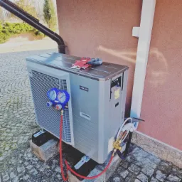 Zewnętrzna jednostka pompy ciepła Nibe AMS 10-8 na kamiennej kostce brukowej, podłączona do instalacji za pomocą przewodów. Na górze leżą robocze rękawice. Widoczne narzędzia pomiarowe.