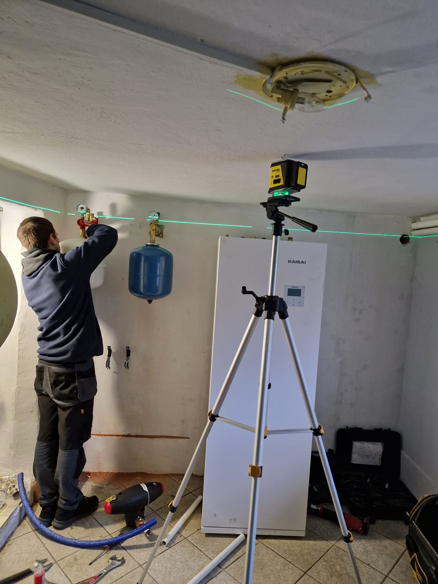 Instalator podłącza elementy instalacji centralnego ogrzewania, widoczny ekspansomat oraz laser krzyżowy na statywie, w tle biała obudowa urządzenia Kaisai.