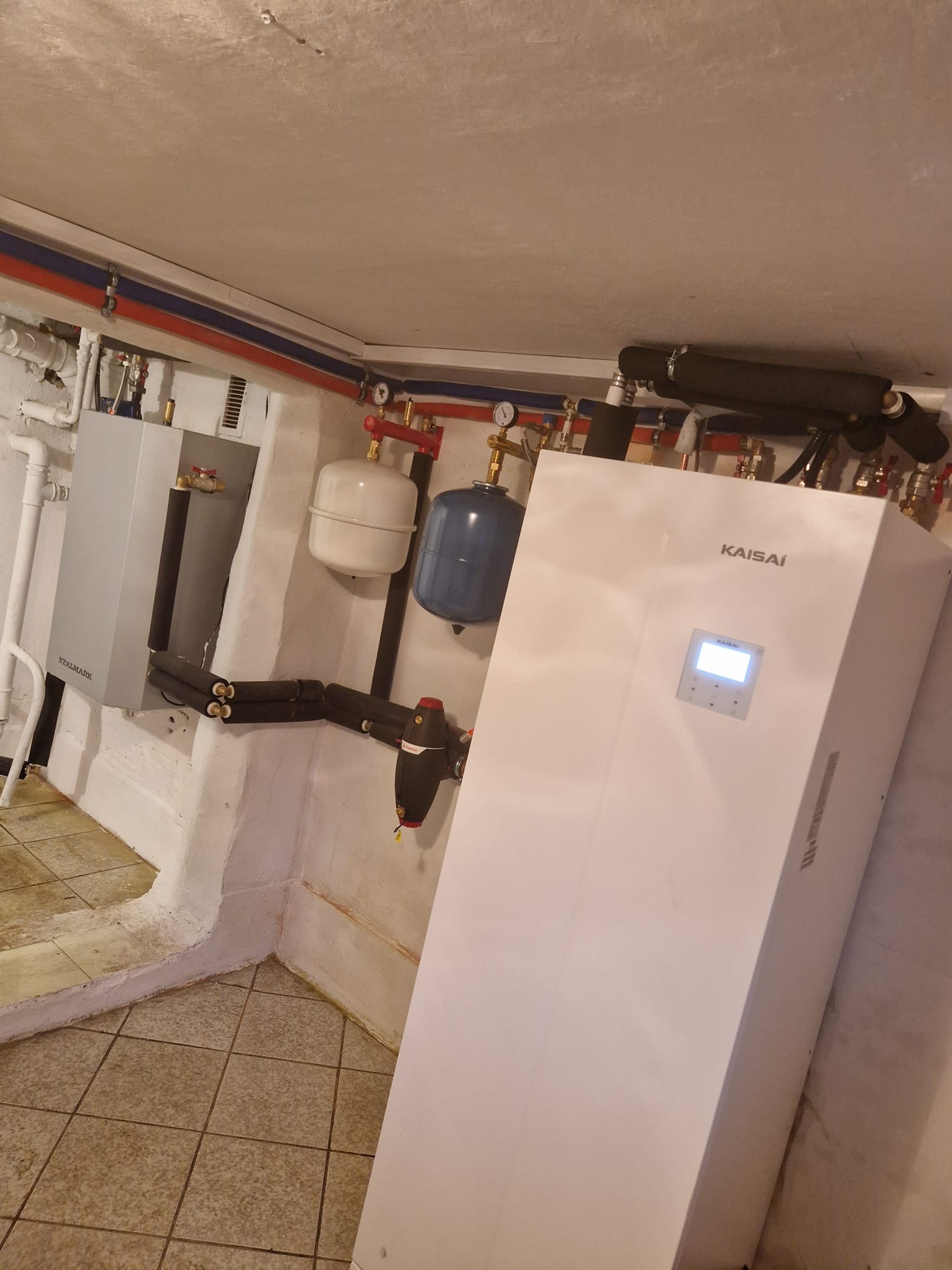 Zainstalowana biała pompa ciepła Kaisai z widocznym panelem sterowania w piwnicy, obok szary zbiornik Stalmark, rury w izolacji i instalacja wodna z manometrami.