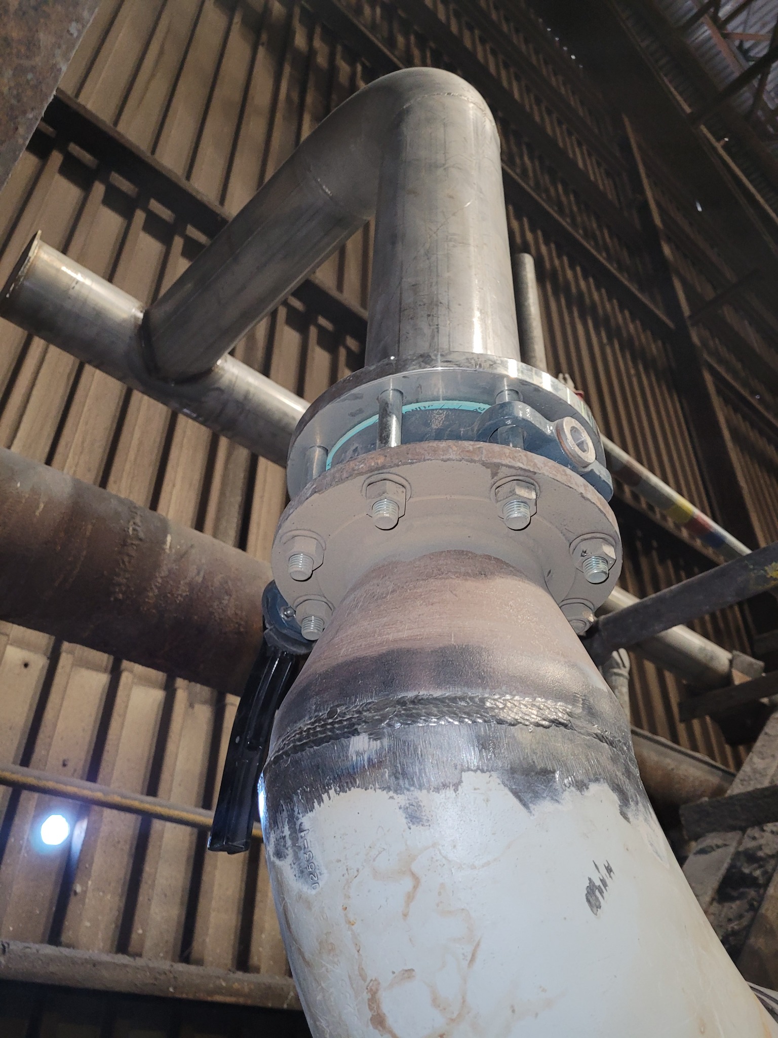 Połączenie spawane rur przemysłowych z kołnierzem skręcanym śrubami, widoczne ślady spawania i malowania, rury prowadzone pod stropem hali.