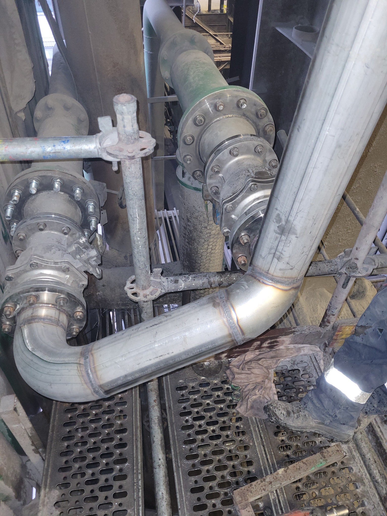 Metalowa rura połączona spawami z kołnierzem, widoczne ślady spawania, w tle inne instalacje przemysłowe i metalowa kratownica. U dołu fragment buta roboczego.