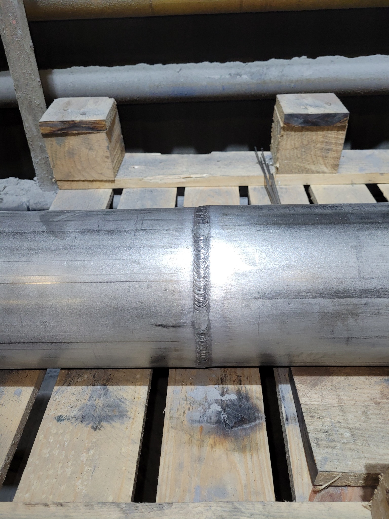 Połączenie spawane na rurze ze stali nierdzewnej, widoczne ślady po spawaniu, ułożone na palecie drewnianej z drewnianymi podkładkami. Widoczne elektrody spawalnicze.