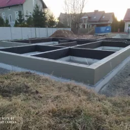 Zalane fundamenty pod budynek z widocznymi ścianami fundamentowymi i izolacją przeciwwilgociową, na działce z piaskiem i trawą, w tle domy jednorodzinne i ogrodzenie.