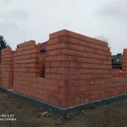 Wznoszone ściany parteru z czerwonej cegły ceramicznej Porotherm, widoczna hydroizolacja fundamentów, w tle domy jednorodzinne i pochmurne niebo.