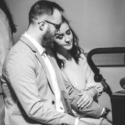 mgartfoto - Czarno-białe zdjęcie pary: mężczyzna w garniturze z brodą i okularach obejmuje kobietę z pierścionkiem, oboje siedzą na krześle obok wózka dziecięcego.