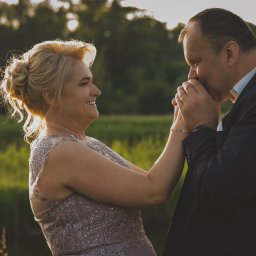 mgartfoto - Elegancka para w plenerze: mężczyzna w granatowym garniturze całuje dłoń uśmiechniętej kobiety w zdobionej sukni, złote światło zachodzącego słońca.