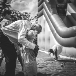 mgartfoto - Czarno-białe zdjęcie przedstawia ojca obejmującego córkę na tle dmuchanego zamku podczas przyjęcia na trawniku, z widoczną inną osobą w tle.