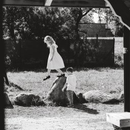 mgartfoto - Czarno-białe zdjęcie przedstawia dziewczynkę w białej sukience skaczącą z kamienia, podczas gdy mały chłopiec w koszuli i jeansach stoi obok, obserwując ją w słoneczny dzień na tle zieleni.