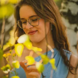 Portret młodej kobiety w okularach, w letniej sukience, delikatnie dotykającej gałązki z żółtymi liśćmi, na tle kory brzozy.