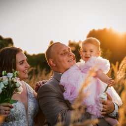 mgartfoto - Szczęśliwa rodzina na tle zachodzącego słońca w polu: mama trzyma bukiet, tata trzyma córeczkę w różowej sukience.