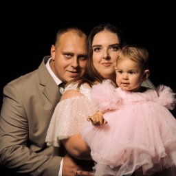 mgartfoto - Portret rodzinny na czarnym tle: ojciec w beżowym garniturze, matka w cekinowej sukni trzyma na rękach córeczkę w różowej tiulowej sukience z perłami.