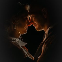 mgartfoto - Intymne zbliżenie pary w ciemności, z delikatnym oświetleniem podkreślającym ich twarze. Kobieta z kolczykami, mężczyzna w garniturze, dotykają się nosami.