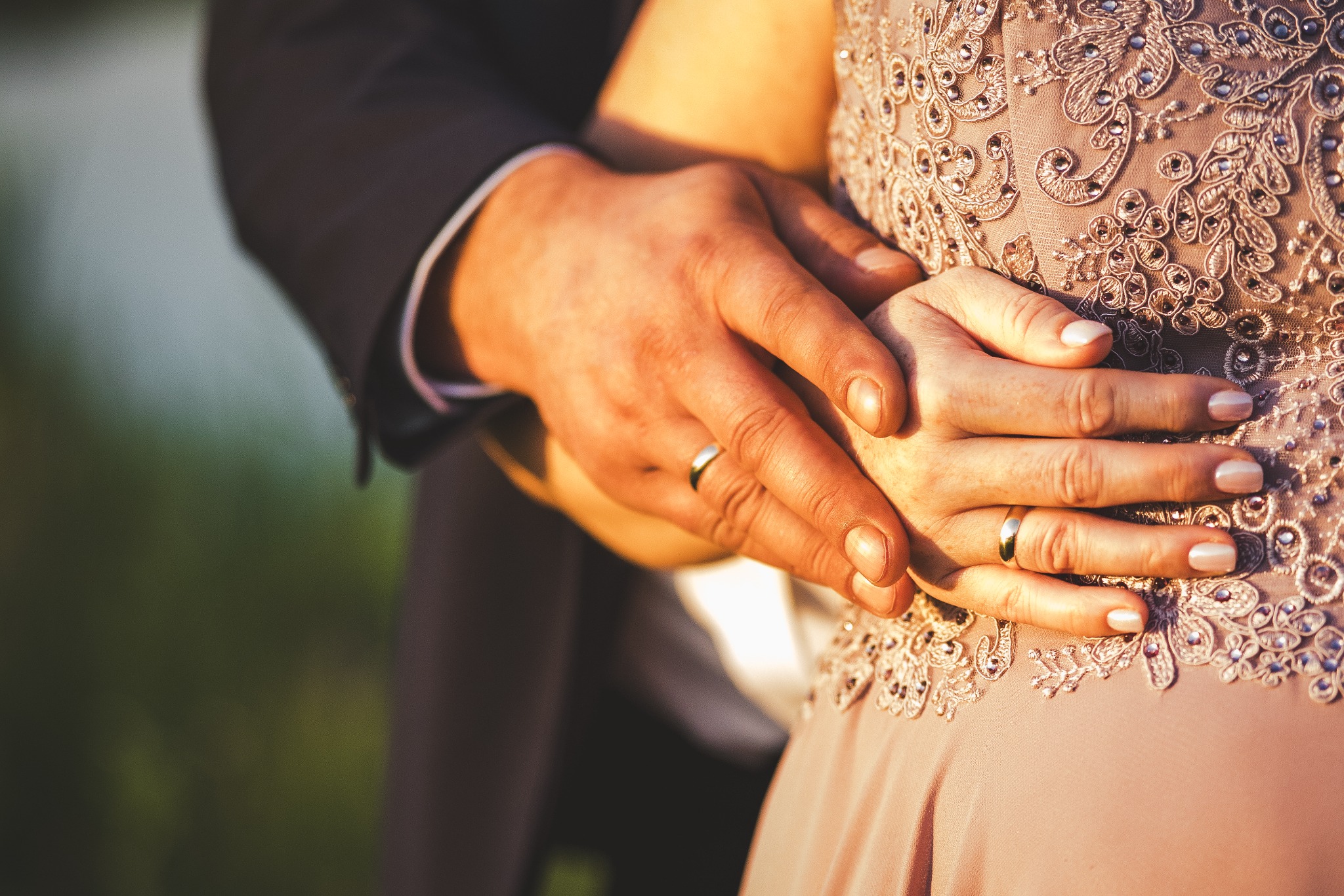 Dłonie pary z obrączkami spoczywają na zdobionej sukni ślubnej w ciepłym oświetleniu.