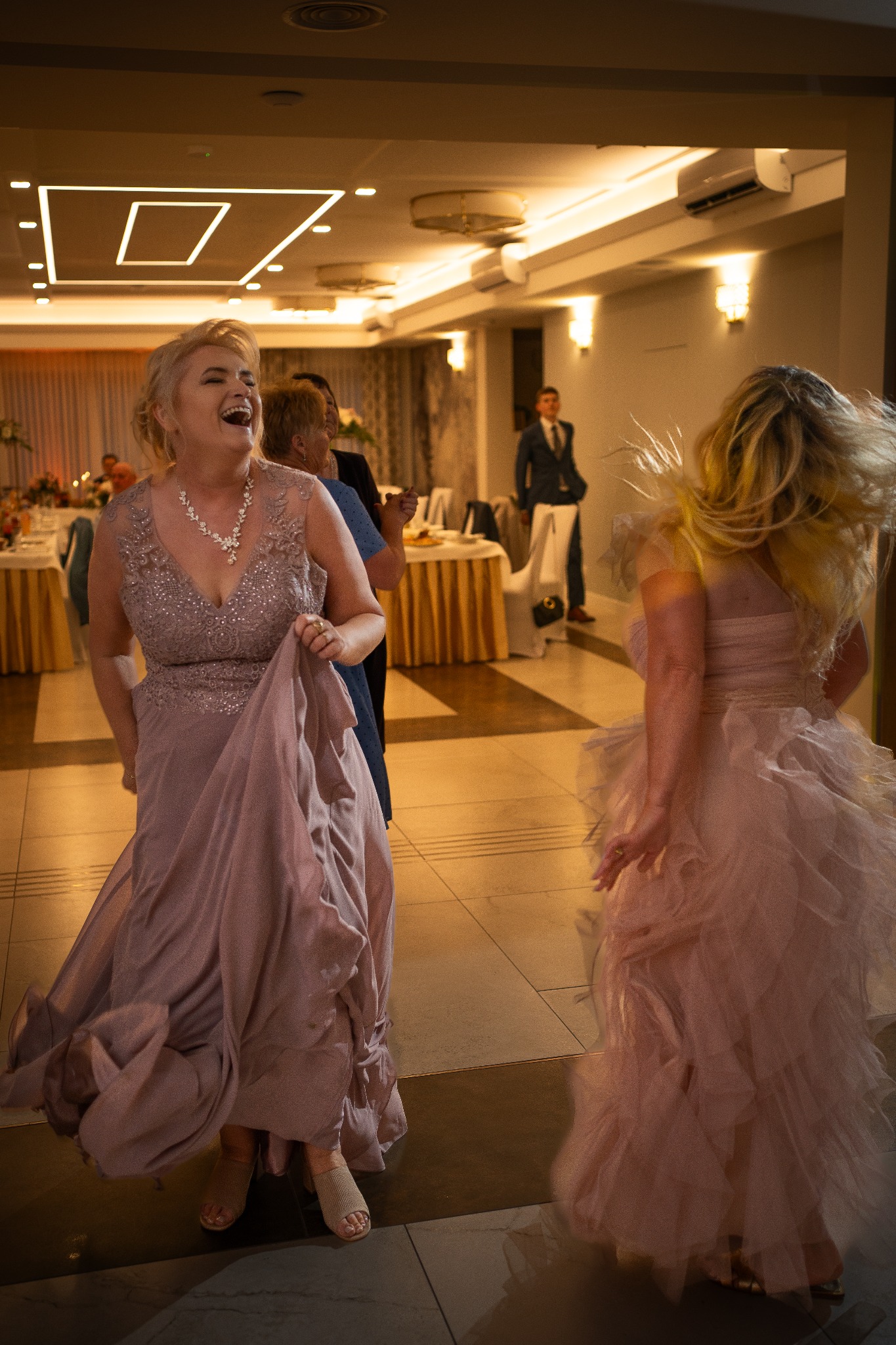 Rozbawiona kobieta w pudrowej sukni tańczy na sali balowej podczas uroczystości, w tle widoczni goście i mężczyzna w garniturze.