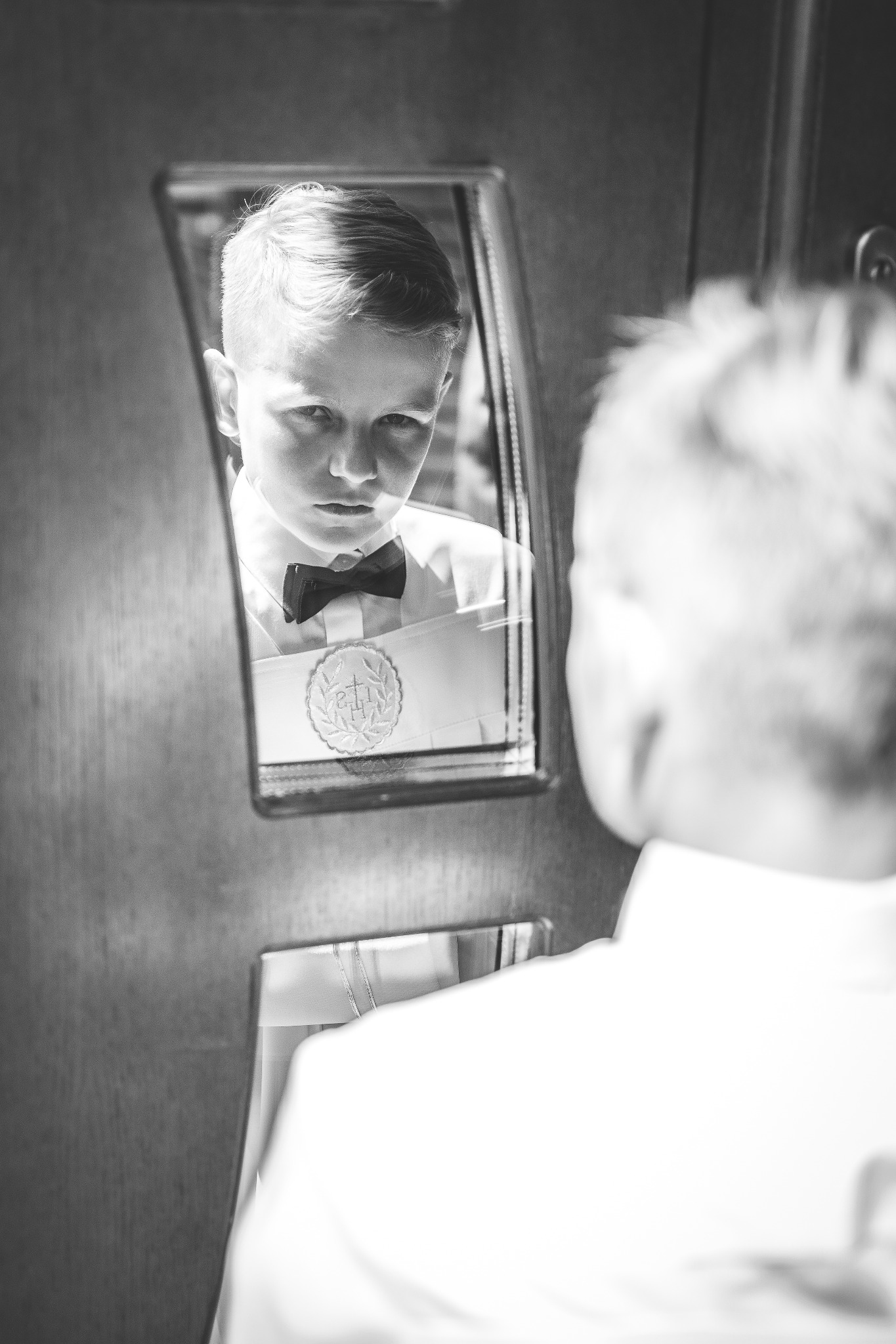 Chłopiec w koszuli i muszce patrzy w lustro, przygotowując się do uroczystości, czarno-biała fotografia.