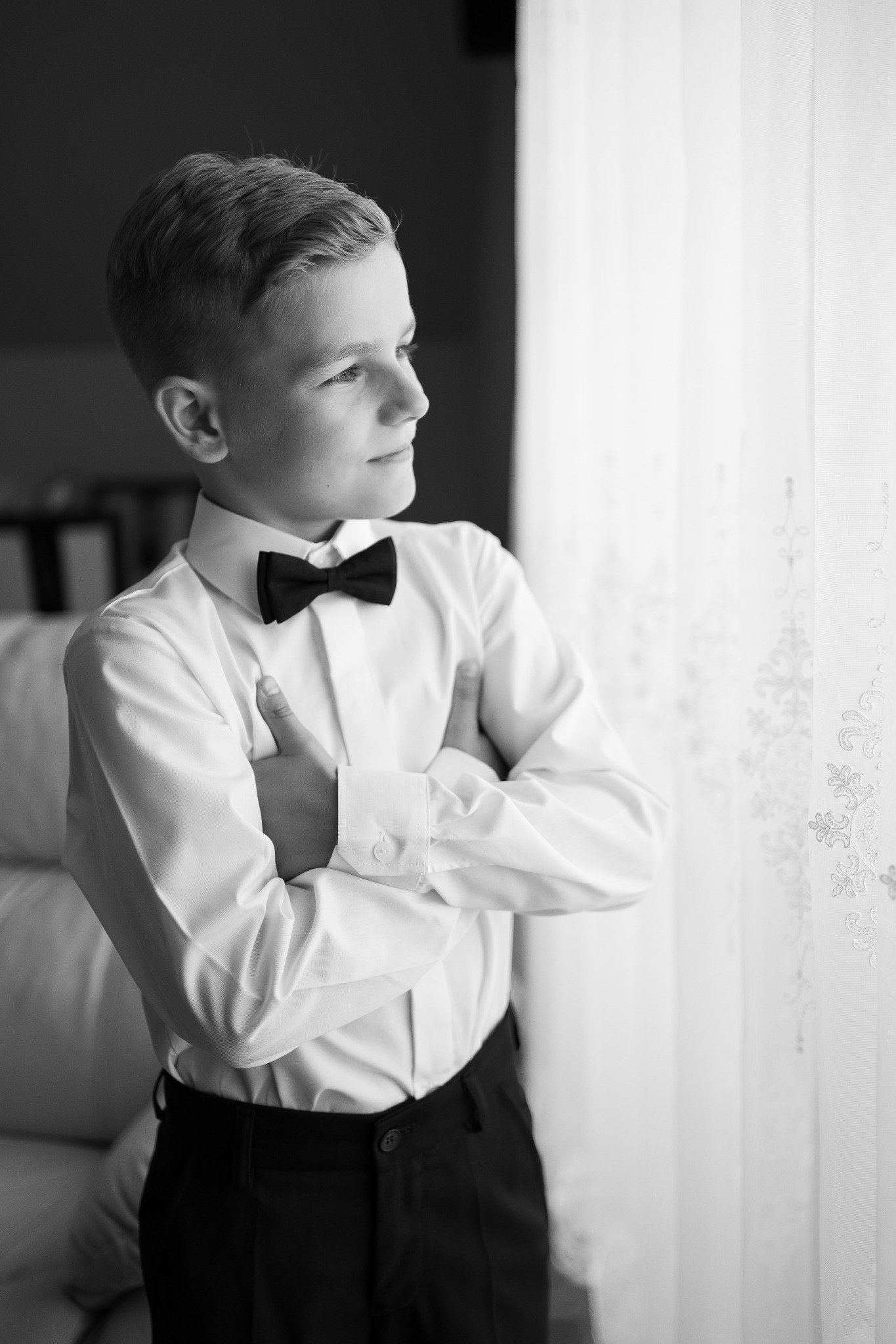 Czarno-białe zdjęcie chłopca w białej koszuli i czarnej muszce, stojącego z założonymi rękami na tle okna z delikatną firanką.