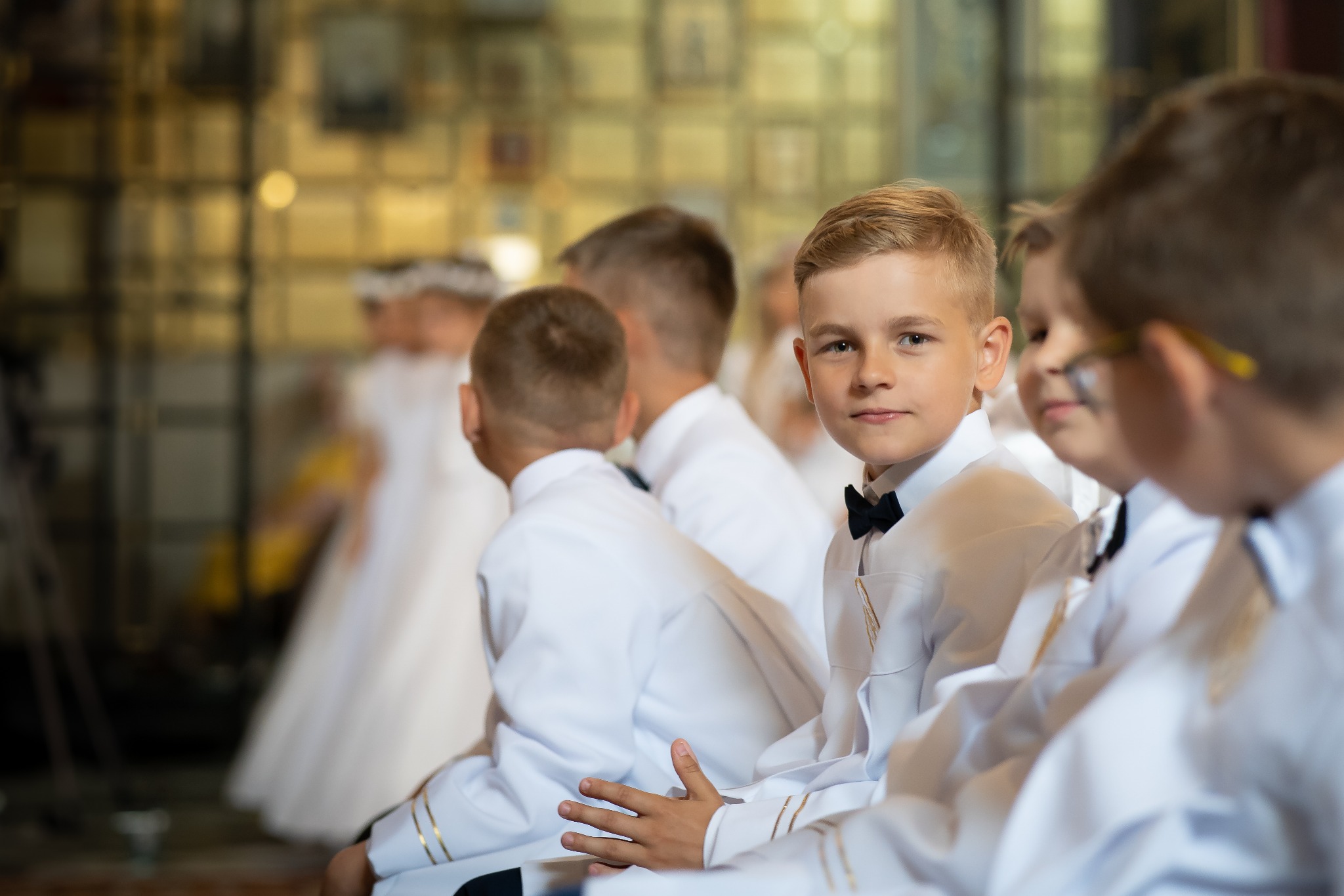 Grupa chłopców w białych strojach komunijnych siedzi w ławkach kościoła, z przodu widoczny chłopiec z blond włosami i czarną muchą, patrzący w obiektyw.