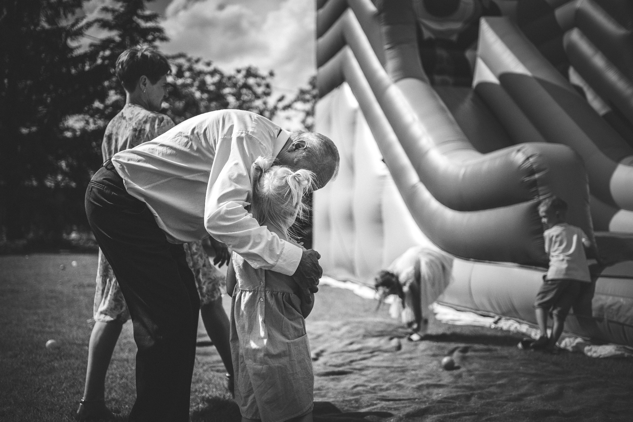 Czarno-białe zdjęcie przedstawia ojca obejmującego córkę na tle dmuchanego zamku podczas przyjęcia na trawniku, z widoczną inną osobą w tle.