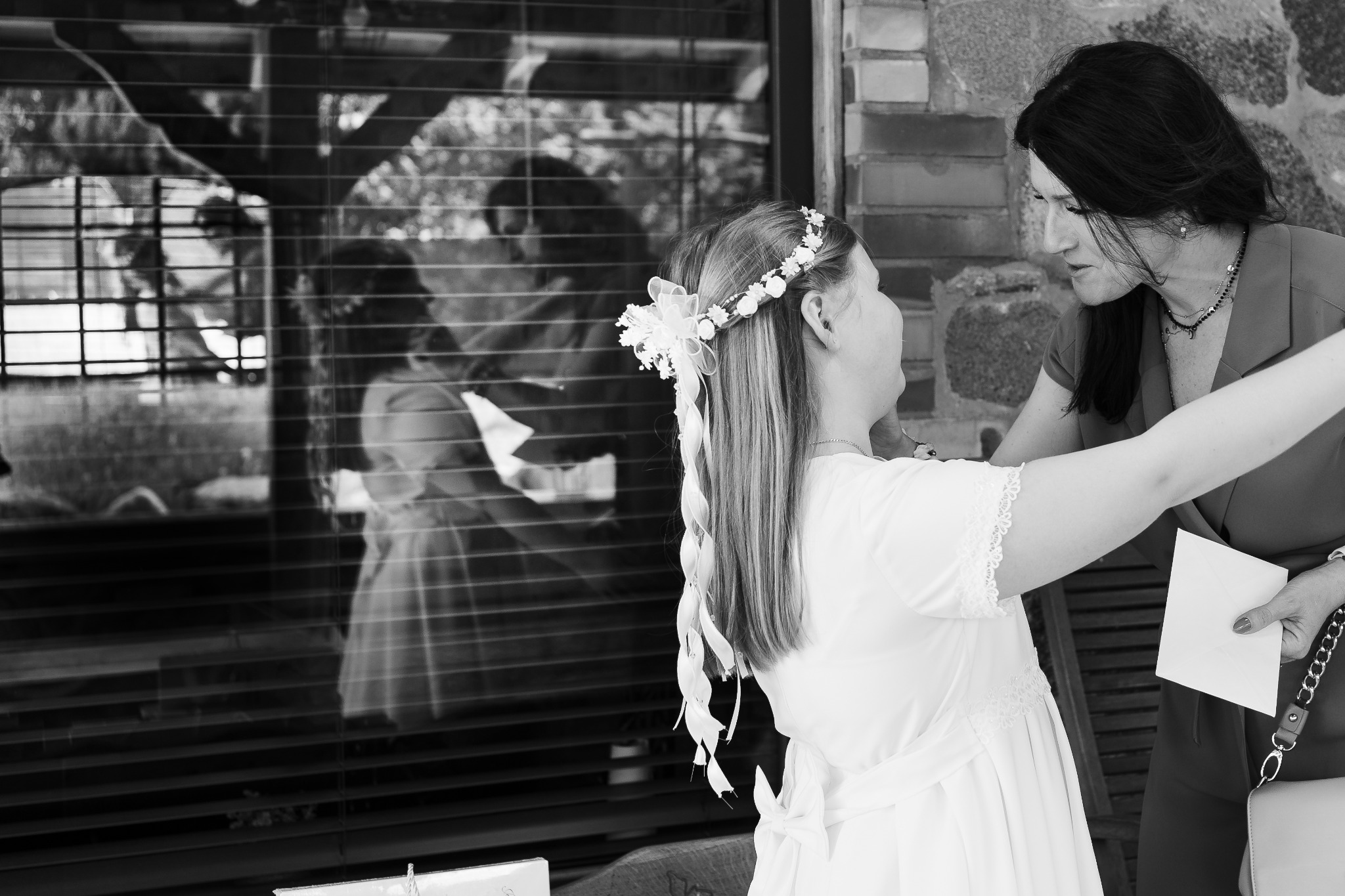 Czarno-białe zdjęcie panny młodej w białej sukni z wiankiem na głowie, poprawianej przez kobietę w ciemnej sukience na tle okna z żaluzjami, w którym odbija się para.