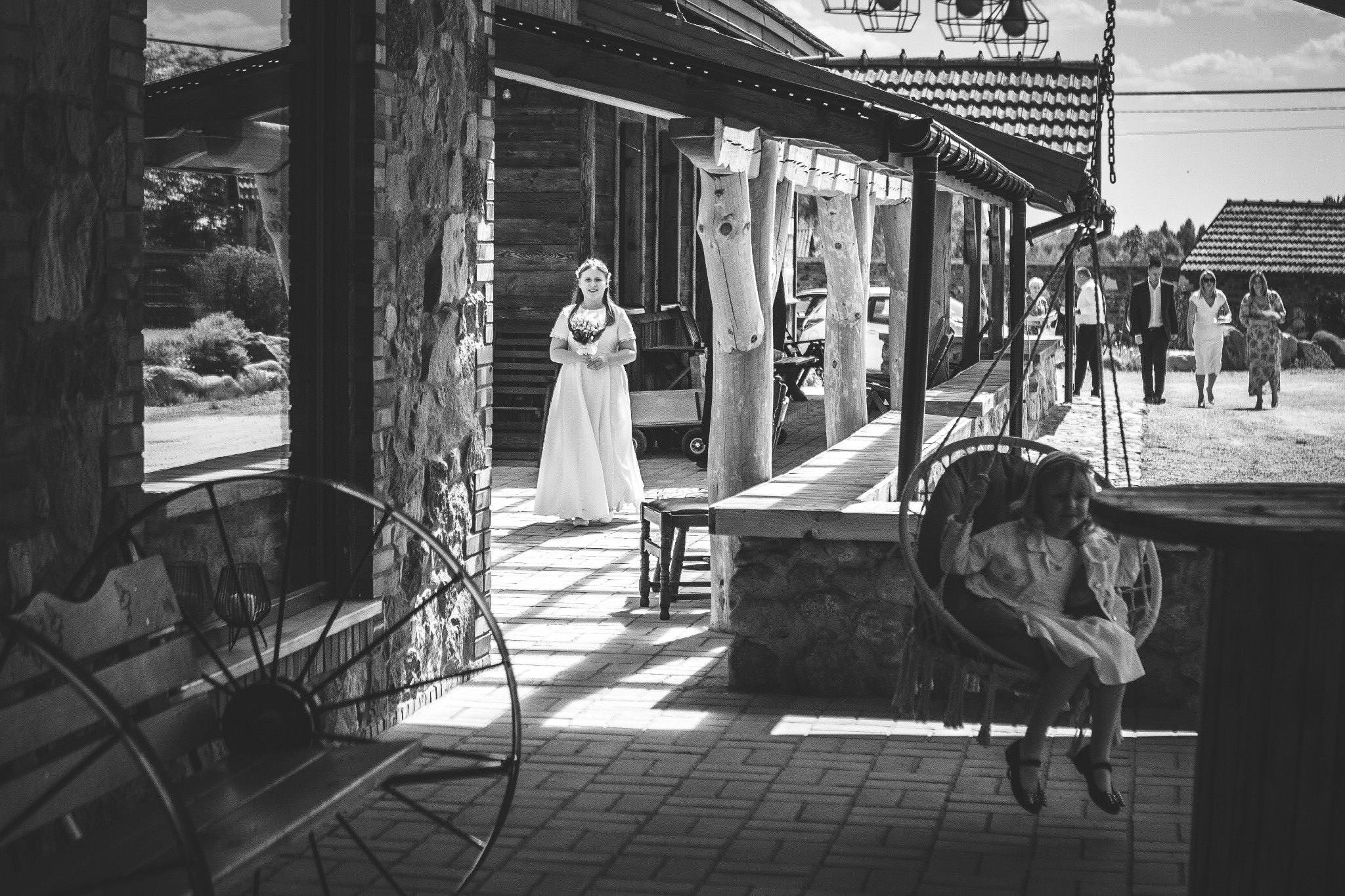 Czarno-białe zdjęcie dziewczynki w białej sukience trzymającej bukiet, stojącej na zewnątrz drewnianej konstrukcji, z dzieckiem siedzącym na huśtawce w pierwszym planie i gośćmi w tle.