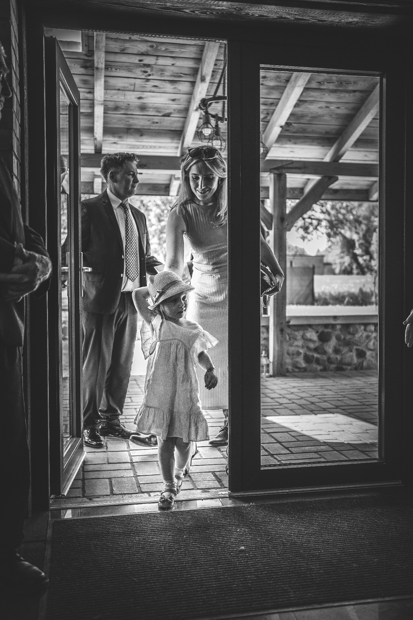 Czarno-białe zdjęcie z wesela: dziewczynka w kapeluszu wchodzi przez otwarte drzwi, za nią uśmiechnięta kobieta i mężczyzna w garniturze, widoczny fragment drewnianej konstrukcji budynku.