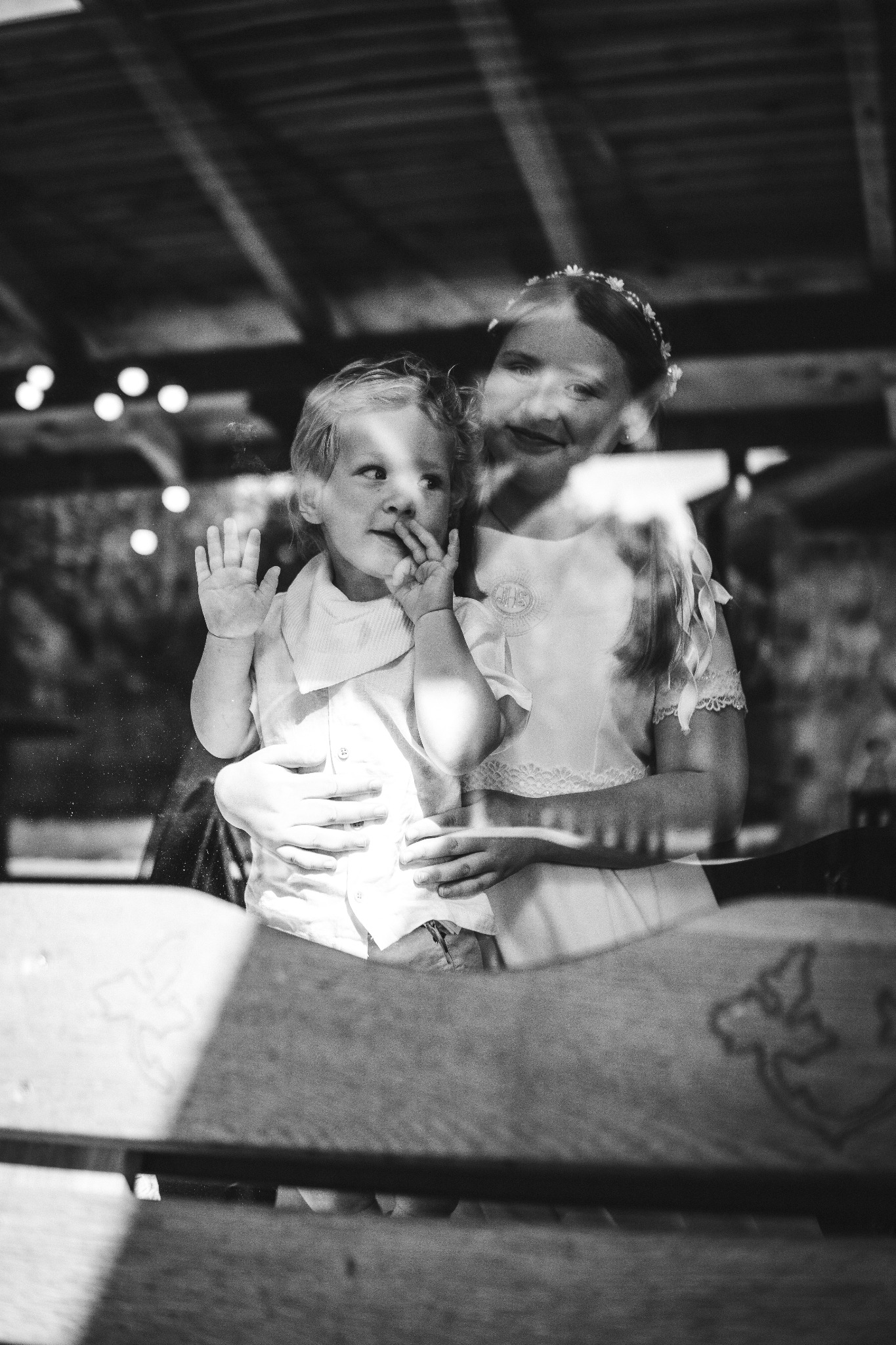 Czarno-białe zdjęcie przedstawia dziewczynkę w sukience komunijnej z wiankiem na głowie, trzymającą na rękach małego chłopca, który przykłada palec do ust i patrzy przez okno. Na pierwszym planie...