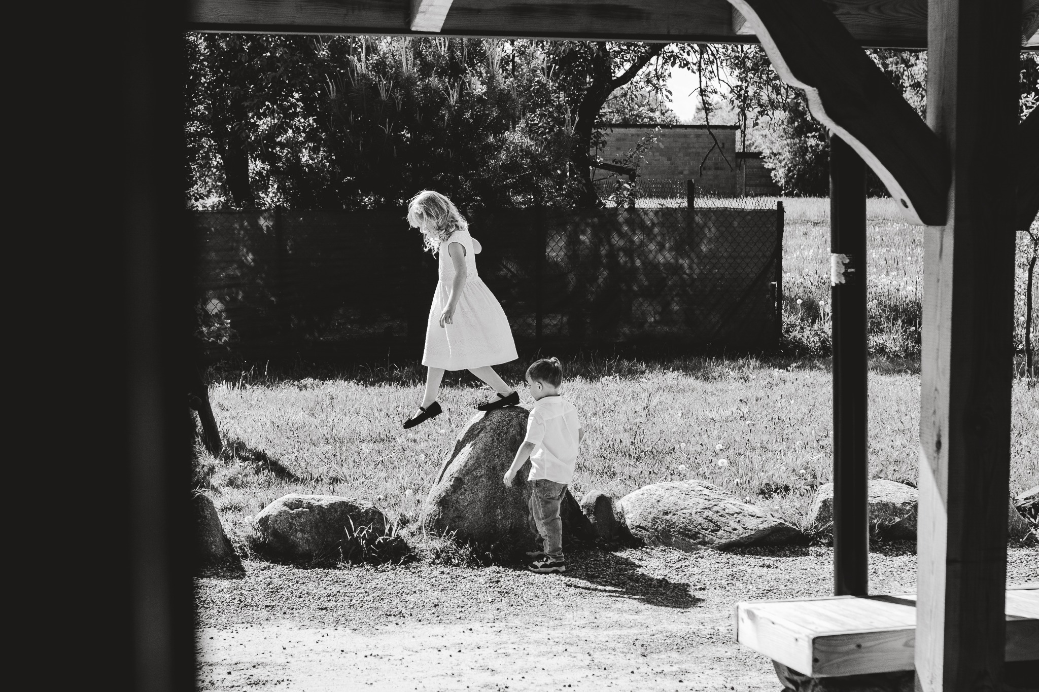 Czarno-białe zdjęcie przedstawia dziewczynkę w białej sukience skaczącą z kamienia, podczas gdy mały chłopiec w koszuli i jeansach stoi obok, obserwując ją w słoneczny dzień na tle zieleni.