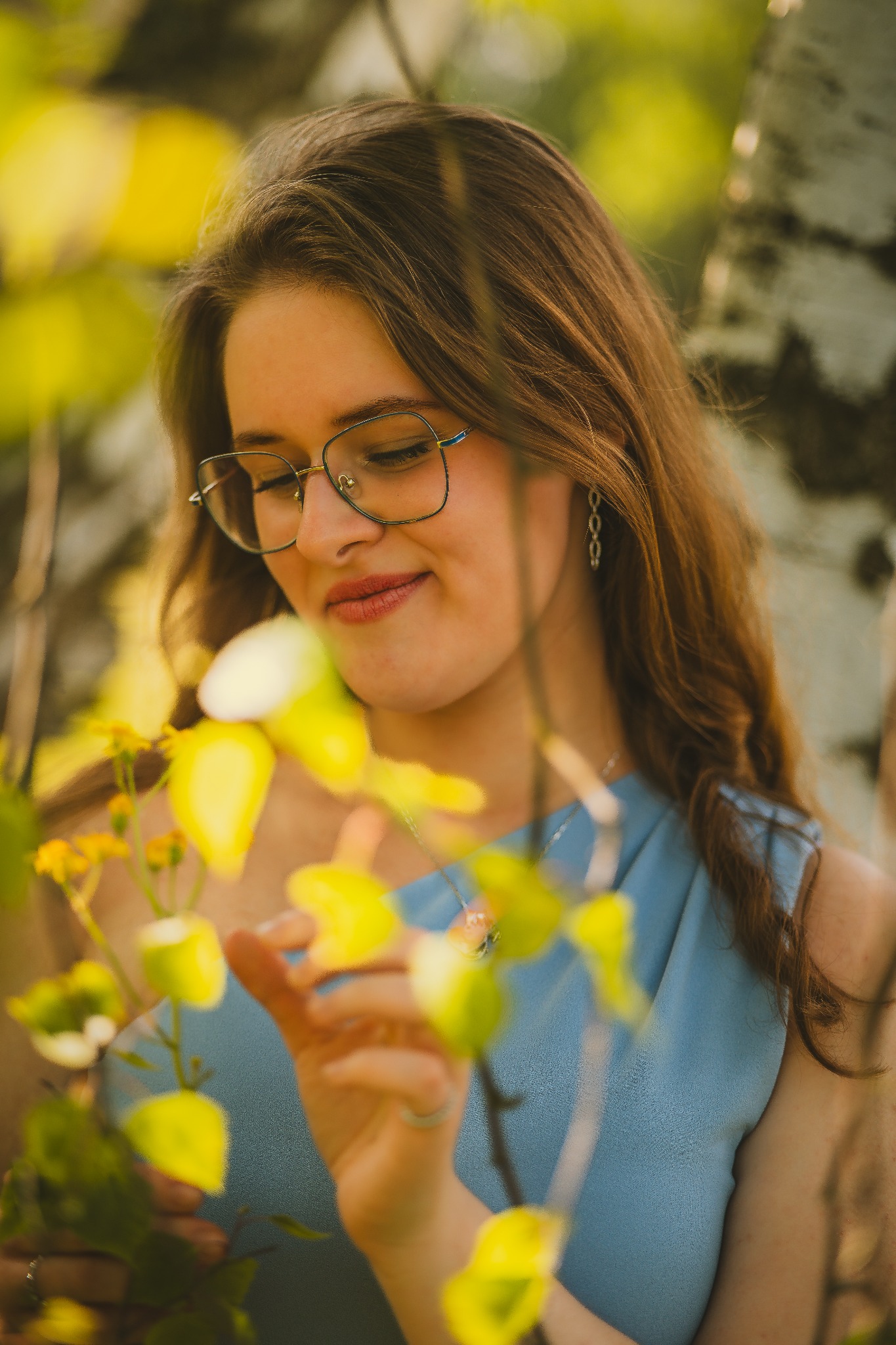 Portret młodej kobiety w okularach, w letniej sukience, delikatnie dotykającej gałązki z żółtymi liśćmi, na tle kory brzozy.