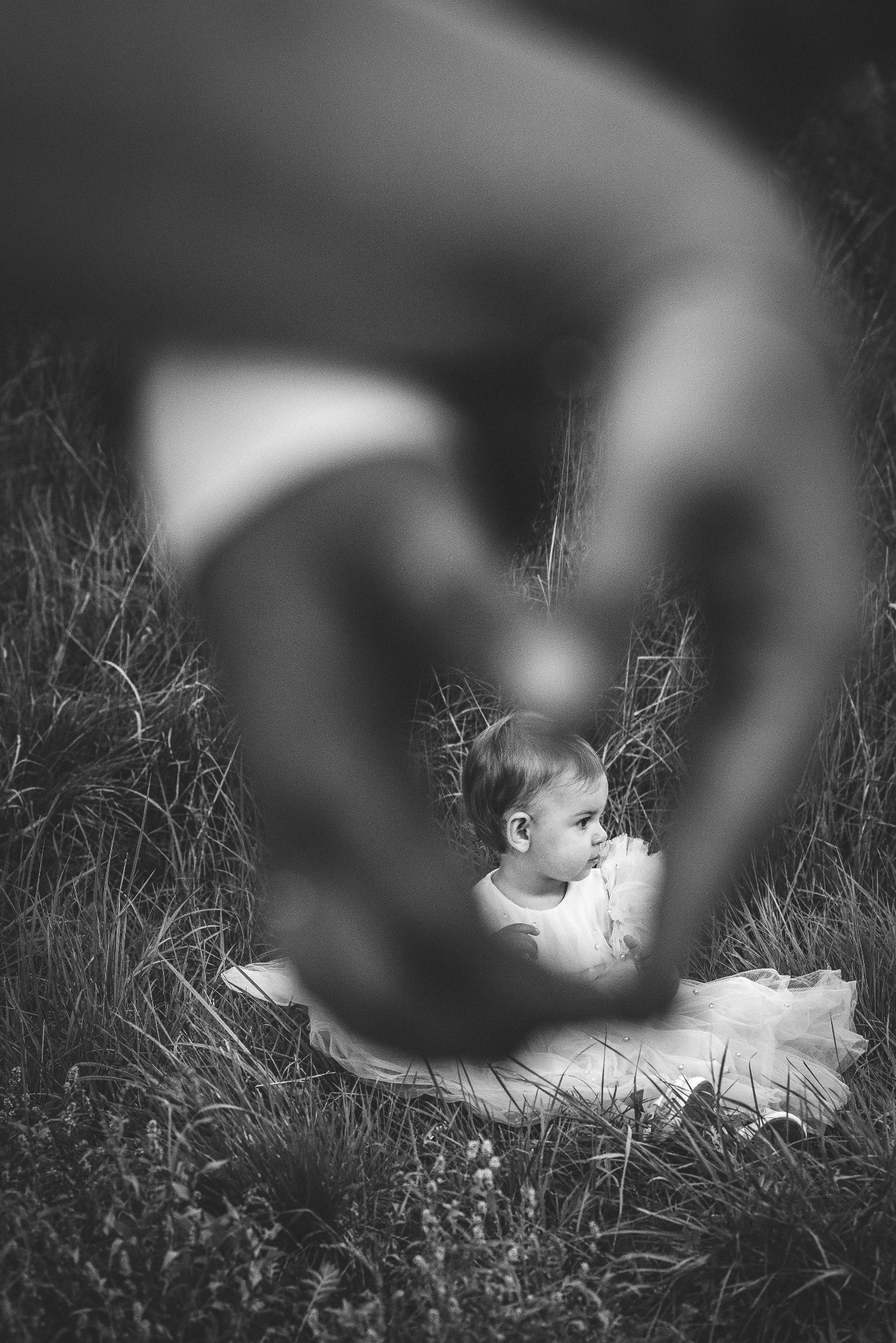 Czarno-białe zdjęcie dziewczynki w białej sukience siedzącej w trawie, uchwyconej przez dłonie tworzące kształt serca w Końskich.