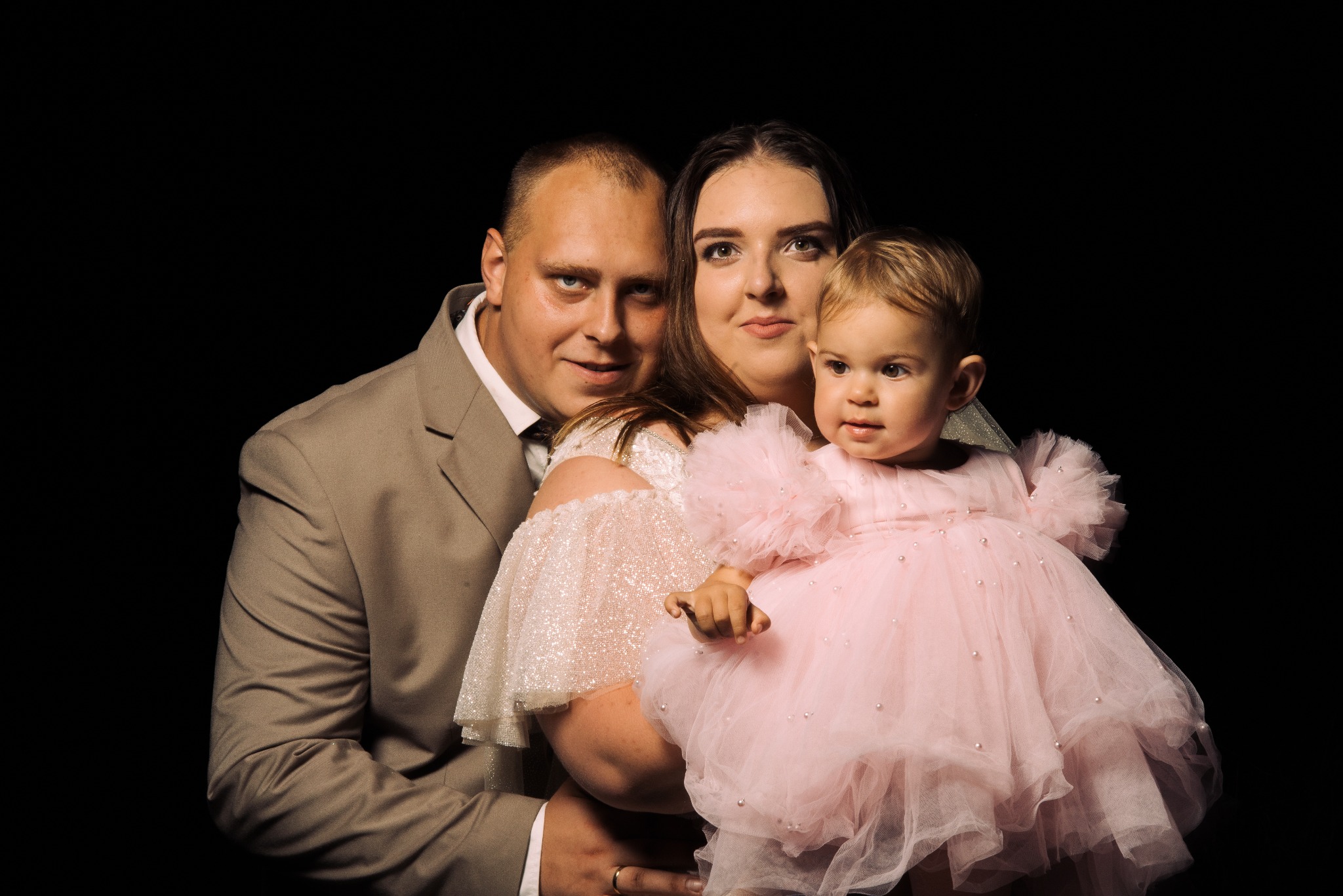 Portret rodzinny na czarnym tle: ojciec w beżowym garniturze, matka w cekinowej sukni trzyma na rękach córeczkę w różowej tiulowej sukience z perłami.