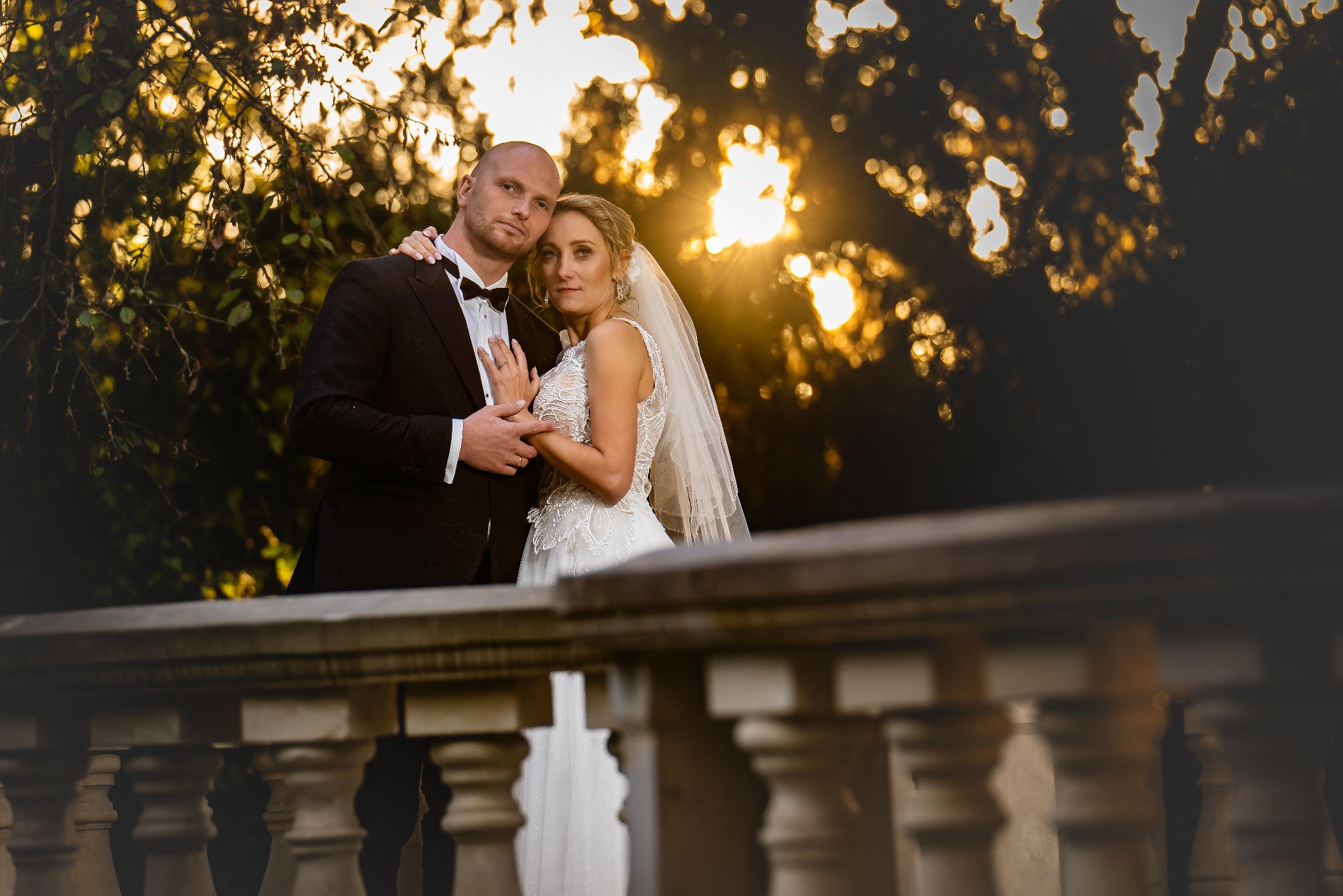 Para młoda w objęciach na tle zachodzącego słońca, widoczna kamienna balustrada. Pan młody w smokingu, panna młoda w koronkowej sukni i welonie.