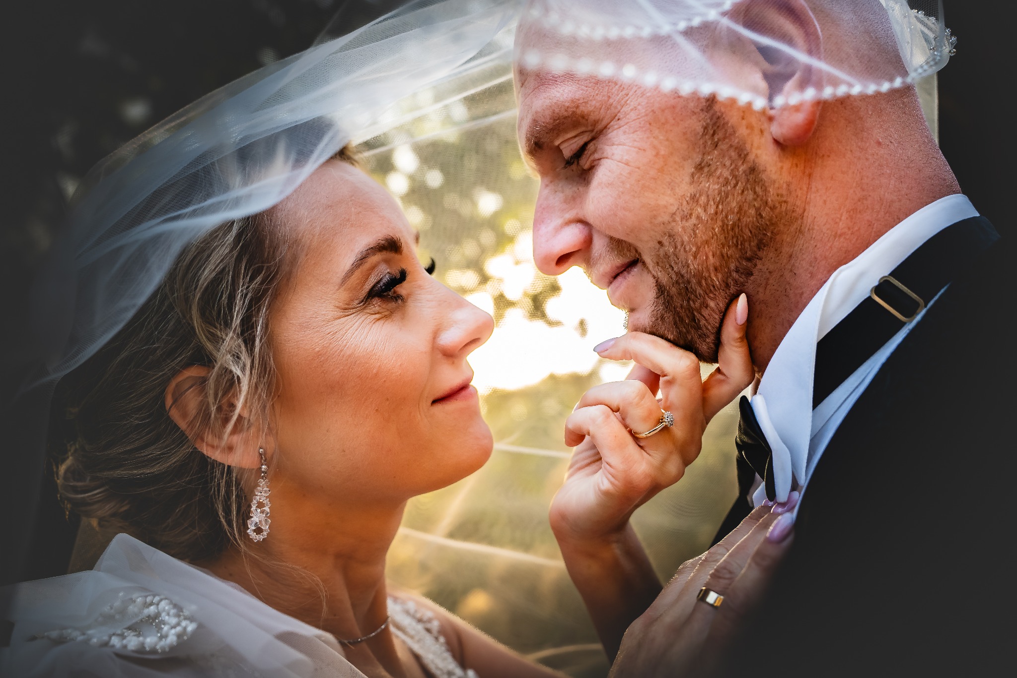 Ujęcie profilowe pary młodej pod welonem, z delikatnym uśmiechem i wzajemnym spojrzeniem, w tle rozmyte drzewa i światło słoneczne. Dłoń panny młodej z pierścionkiem.