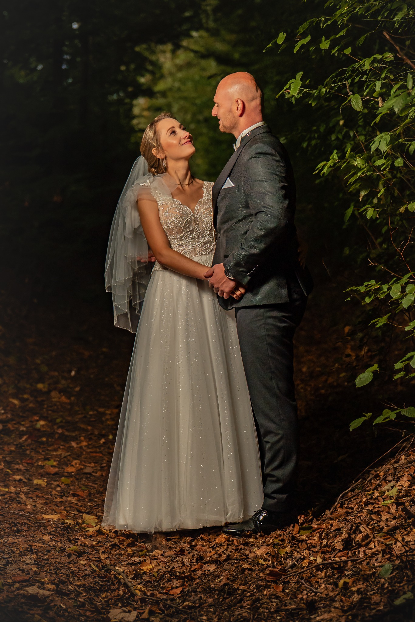 Para młoda w leśnej scenerii: panna młoda w koronkowej sukni spogląda na pana młodego w eleganckim garniturze. Romantyczna atmosfera, naturalne światło.