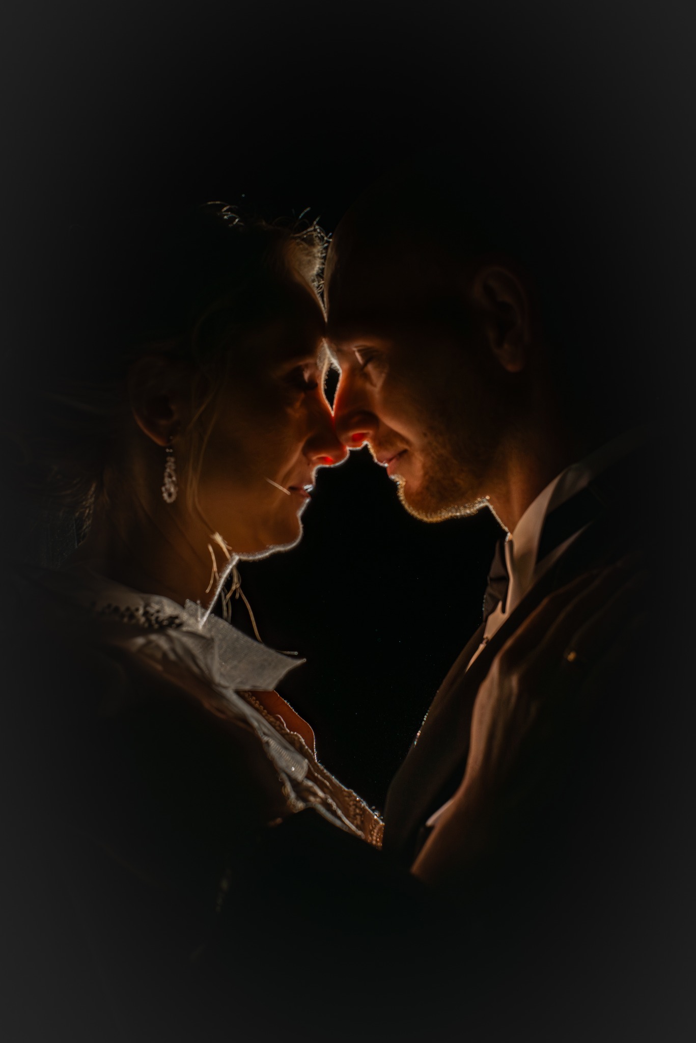 Intymne zbliżenie pary w ciemności, z delikatnym oświetleniem podkreślającym ich twarze. Kobieta z kolczykami, mężczyzna w garniturze, dotykają się nosami.