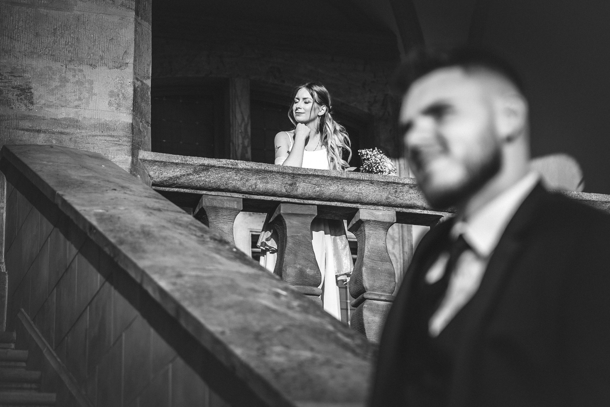 Czarno-białe zdjęcie panny młodej z bukietem na balkonie, z rozmazaną sylwetką mężczyzny w garniturze na pierwszym planie. Ujęcie z dołu, podkreślające architekturę.