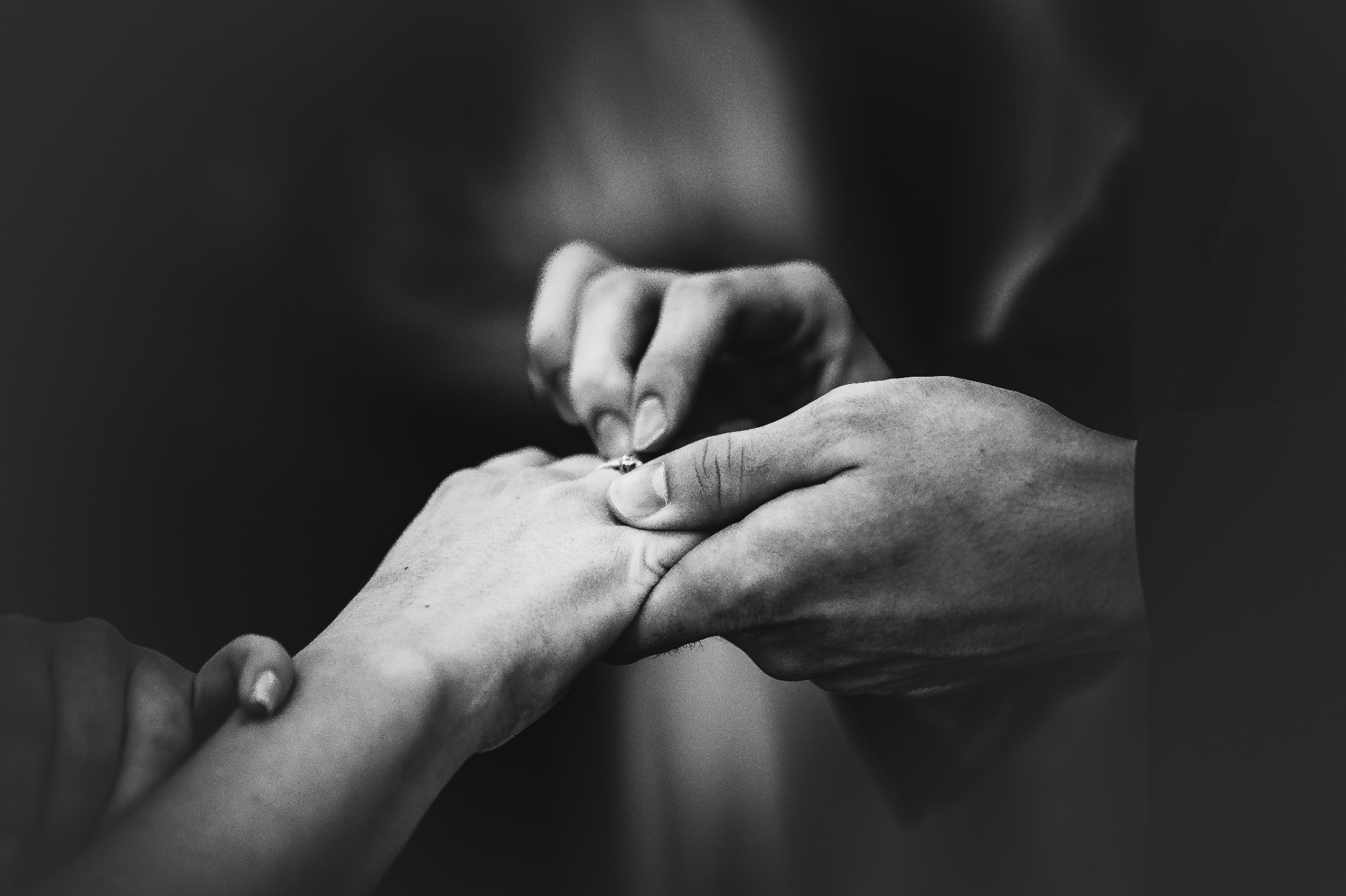 Czarno-białe zbliżenie dłoni podczas zaręczyn: mężczyzna zakłada pierścionek na palec kobiety. Delikatne światło podkreśla moment.