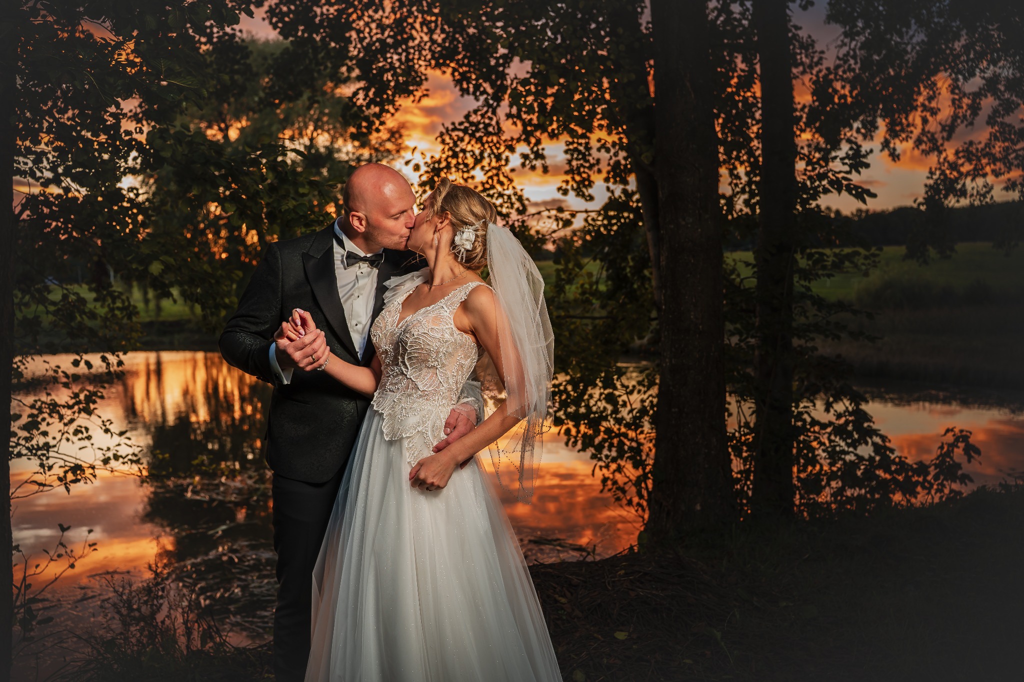 Pocałunek pary młodej na tle jeziora o zachodzie słońca. Pan młody w smokingu, panna młoda w koronkowej sukni i welonie. Romantyczna sceneria z drzewami.