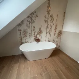 Biała, wolnostojąca wanna na drewnopodobnej podłodze w łazience na poddaszu, ściana z motywem roślinnym, widok z niskiej perspektywy.