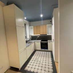 Brulab Spółka z ograniczona odpowiedzialnością - Jasna kuchnia z niebieskim sufitem i mozaikową podłogą. Widoczne białe szafki, czarny blat i sprzęt AGD. Podłoga zabezpieczona kartonem. Nietypowe połączenie kolorów.