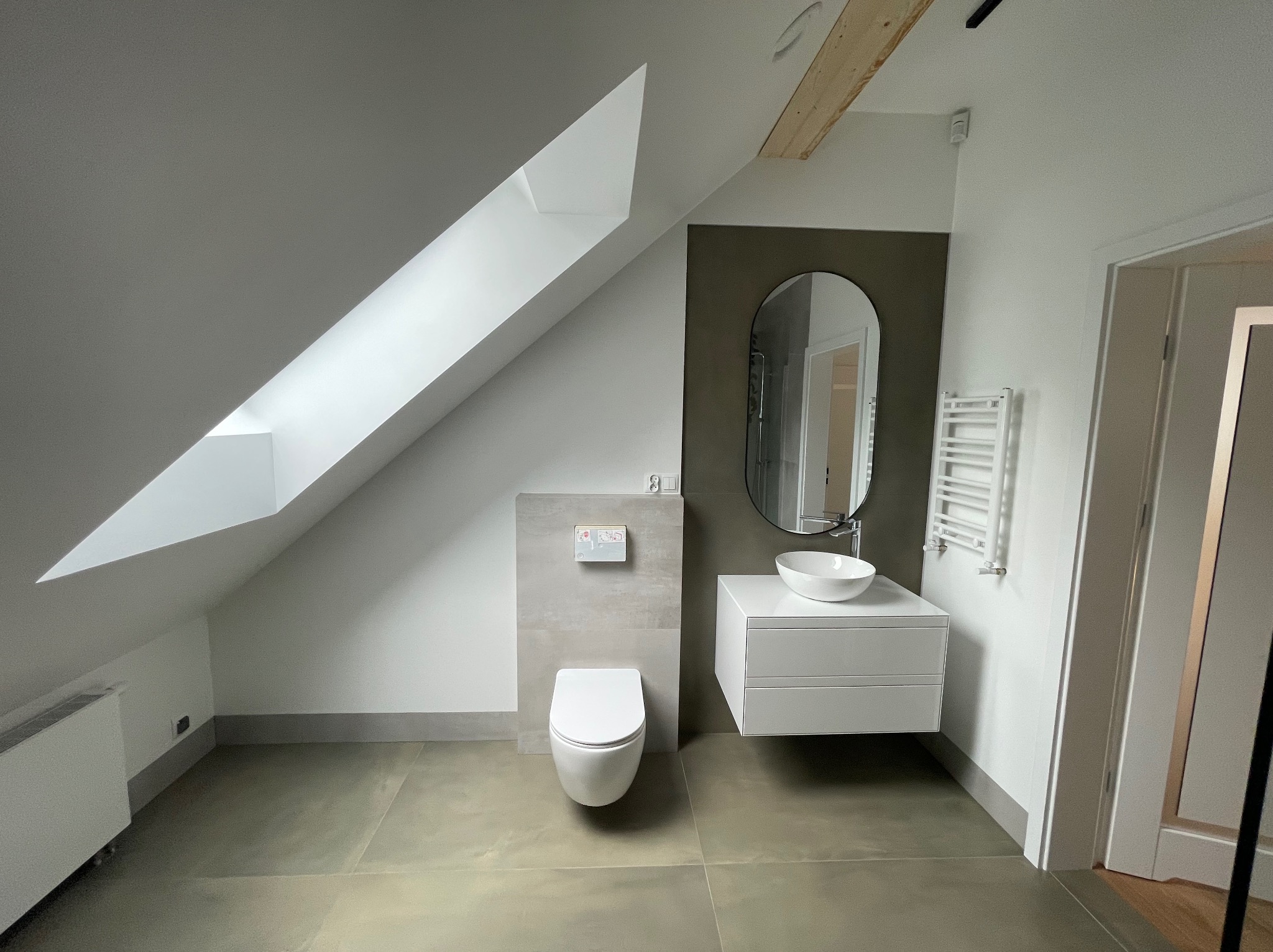 Nowoczesna łazienka na poddaszu z oknem dachowym, umywalką nablatową, wiszącą toaletą i grzejnikiem drabinkowym. Szaro-biała kolorystyka, minimalistyczny design.