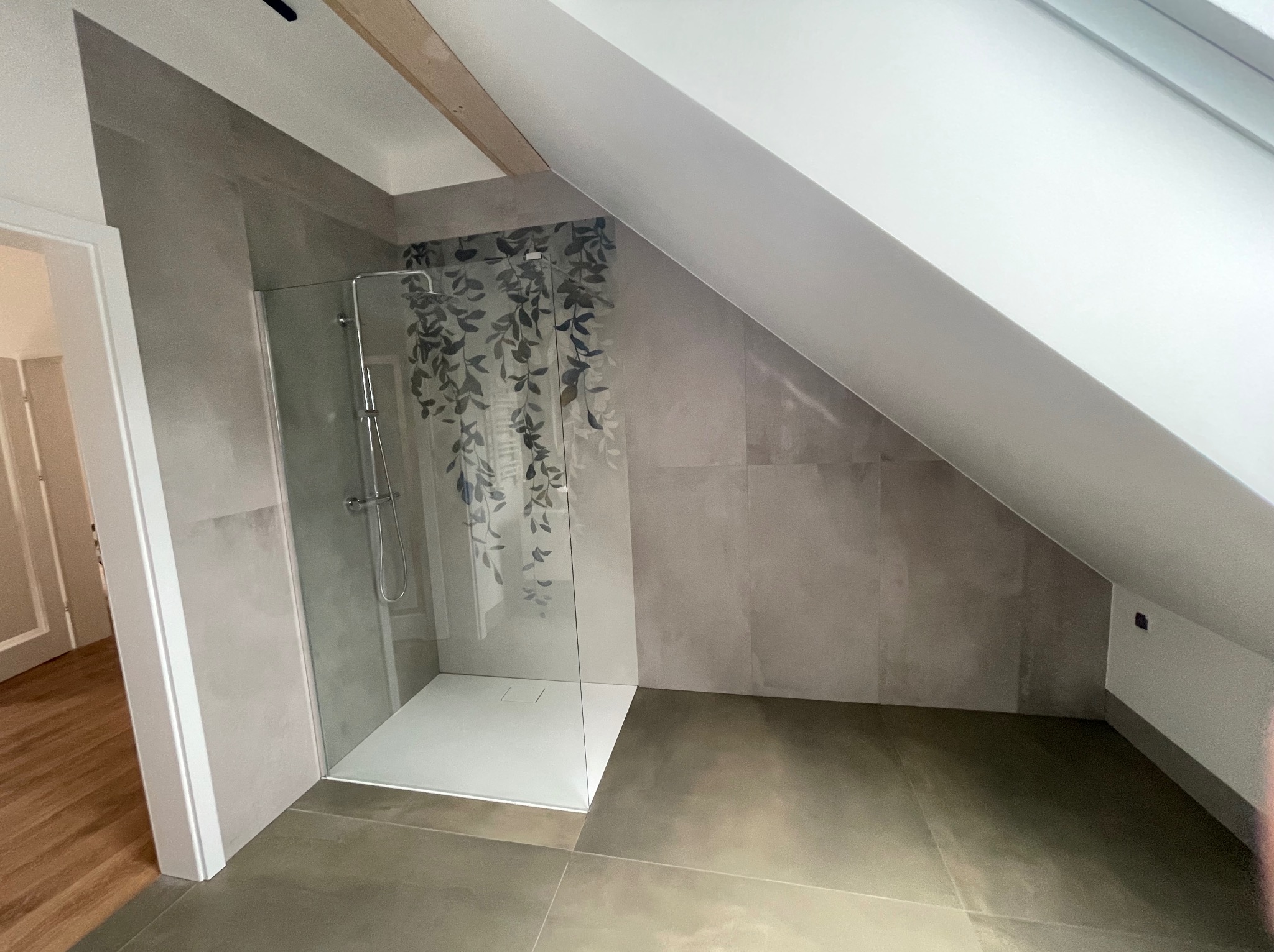 Wykończona łazienka na poddaszu ze szklanym prysznicem z motywem roślinnym. Szare płytki na ścianach i podłodze, biały brodzik. Widoczny fragment drewnianej belki stropowej.