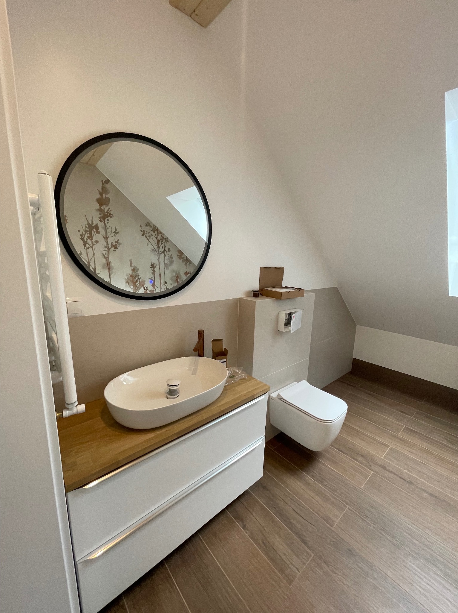 Nowoczesna łazienka na poddaszu z drewnopodobną podłogą, owalną umywalką na drewnianym blacie i okrągłym lustrem z widocznym wzorem na ścianie.