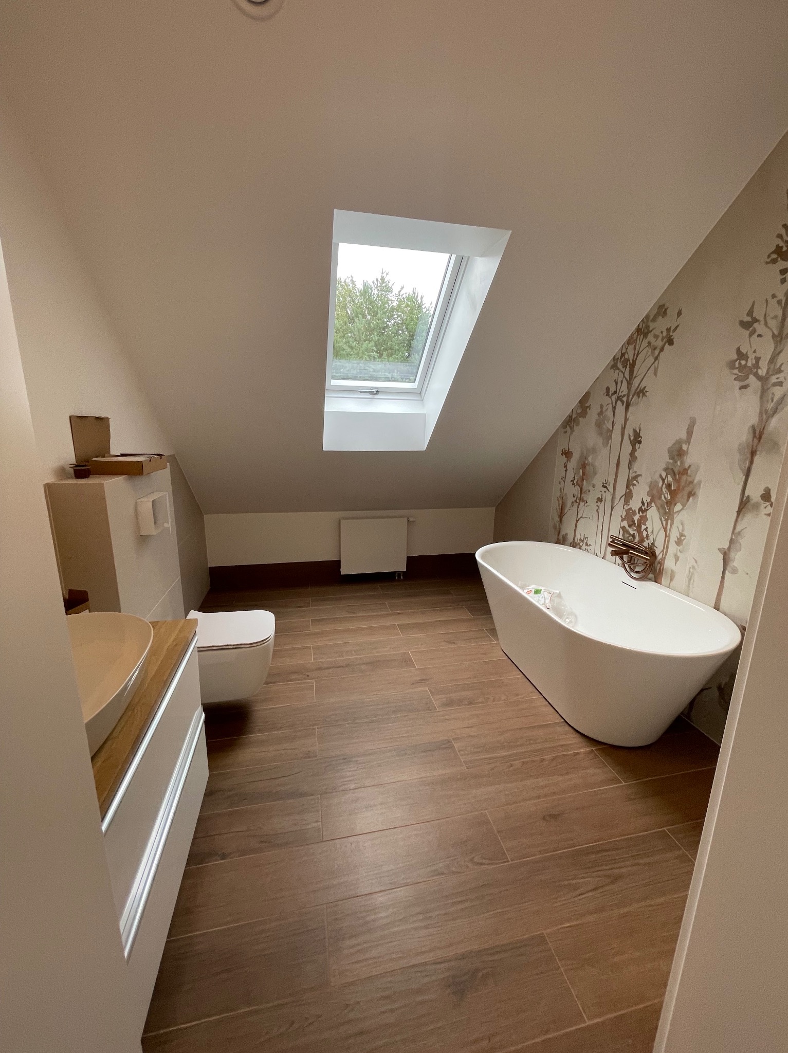 Jasna łazienka na poddaszu z oknem dachowym, drewnopodobną podłogą i wolnostojącą wanną na tle ściany z motywem roślinnym. Widoczna umywalka i toaleta.