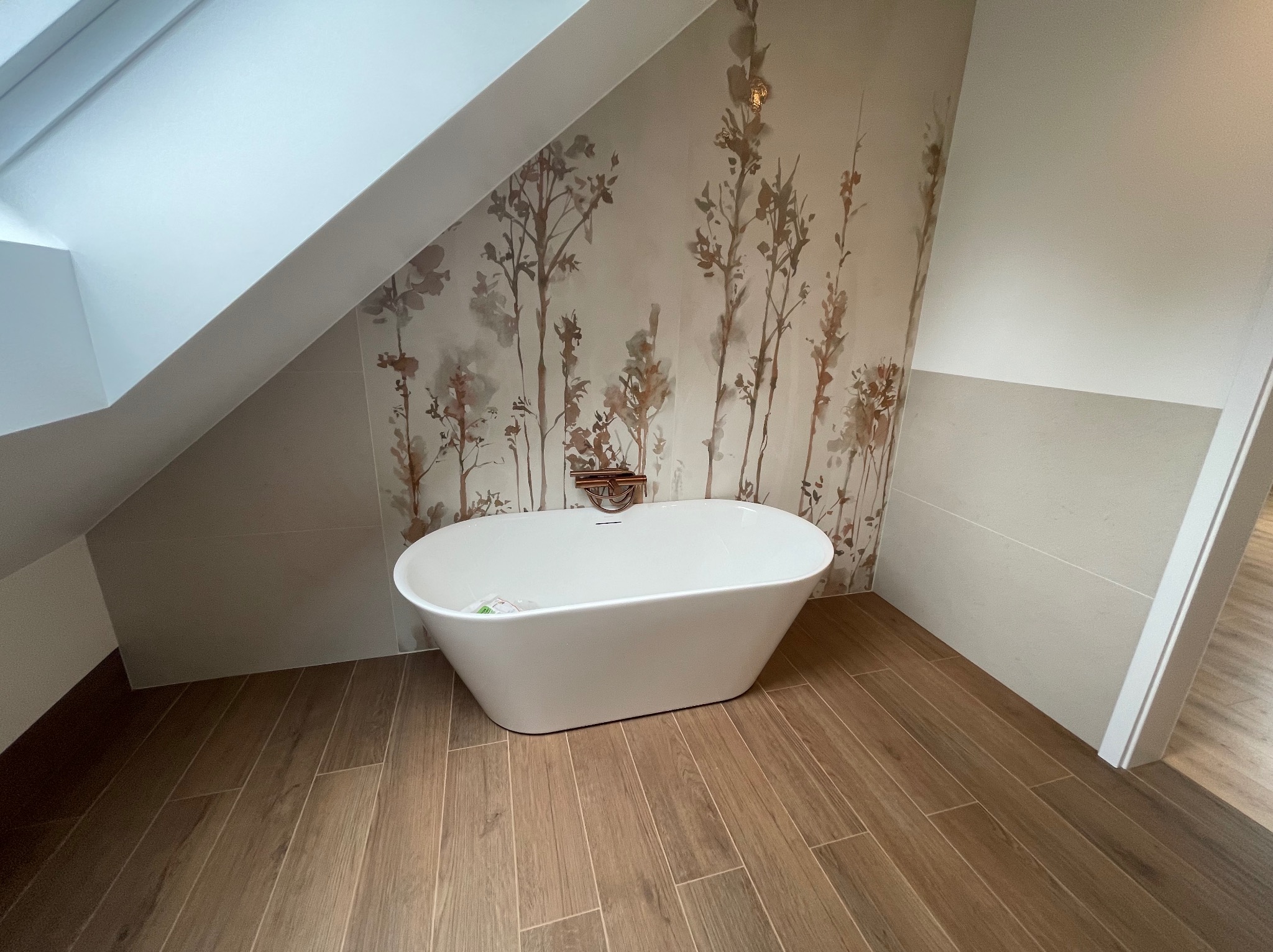 Biała, wolnostojąca wanna na drewnopodobnej podłodze w łazience na poddaszu, ściana z motywem roślinnym, widok z niskiej perspektywy.