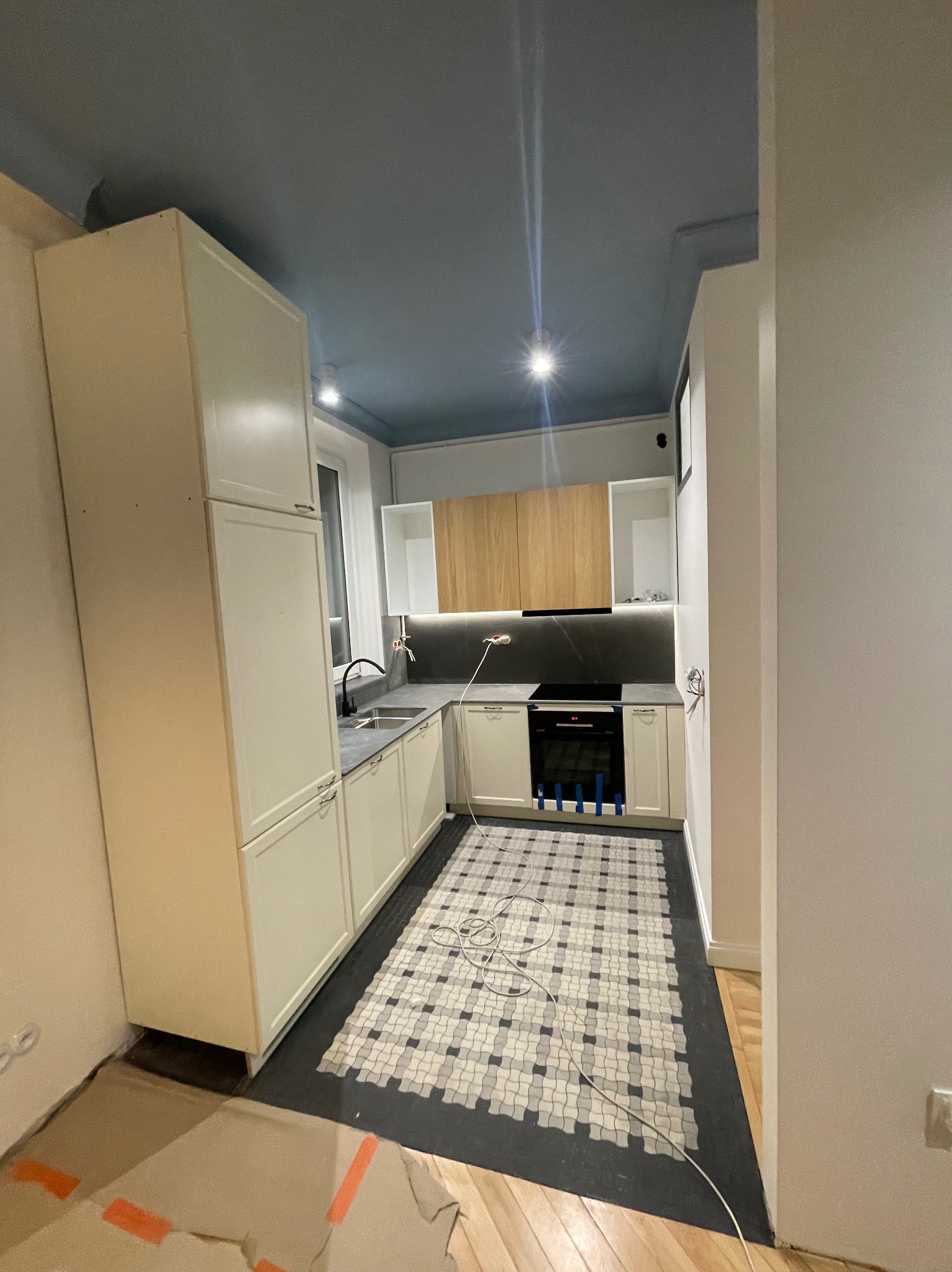 Jasna kuchnia z niebieskim sufitem i mozaikową podłogą. Widoczne białe szafki, czarny blat i sprzęt AGD. Podłoga zabezpieczona kartonem. Nietypowe połączenie kolorów.