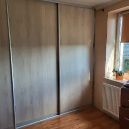 Jasna, drewnopodobna szafa przesuwna z aluminiowymi ramami, widoczna w pokoju z parkietem, grzejnikiem i rośliną doniczkową na parapecie okna