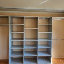 Krystian Szkotak - Szara, otwarta szafa wnękowa z przesuwnymi drzwiami, podzielona na równe kwadratowe i prostokątne półki, zamontowana w pokoju z beżowymi ścianami i drewnianą podłogą. Widoczne listwy dekoracyjne...