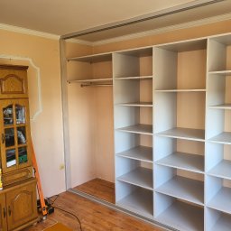 Krystian Szkotak - Wnętrze szafy wnękowej z białymi półkami, widoczny drążek na ubrania, obok stoi drewniany kredens i narzędzia pracy.